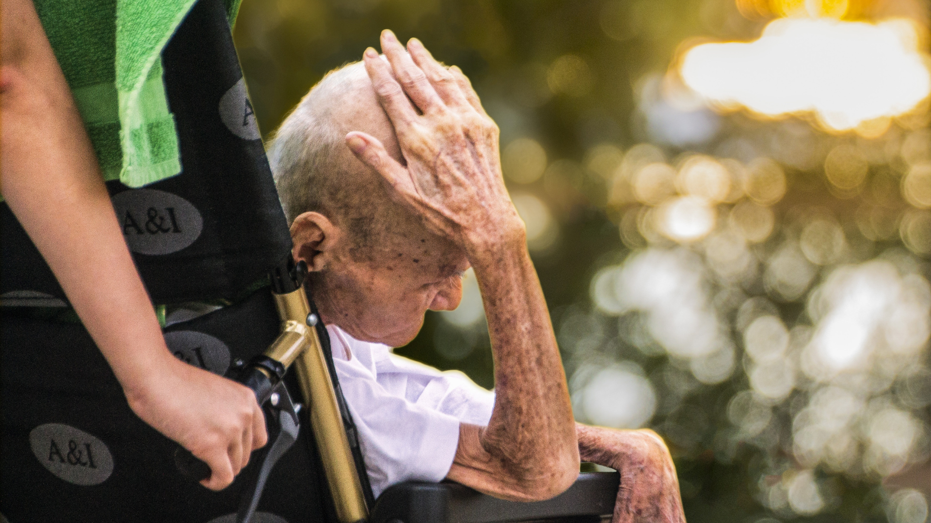 Personne âgée en fauteuil roulant accompagnée à l’extérieur, main posée sur la tête.