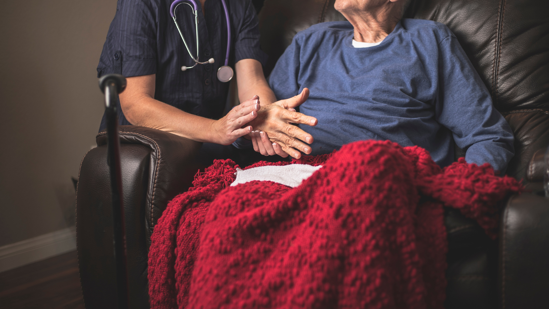 Infirmière à domicile tenant la main d’un patient âgé assis sur un canapé, couverture rouge.